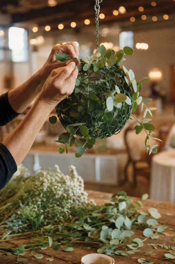 Hands inserting eucalyptus into a suspended floral cage during an installation build