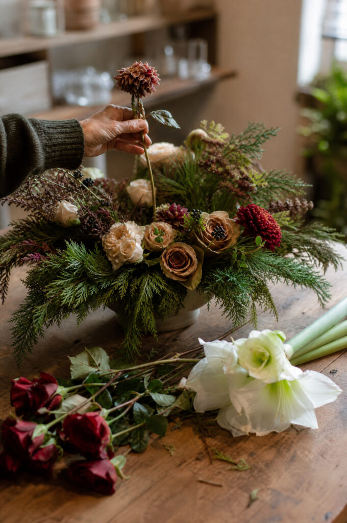 Hand swapping a spent dahlia for fresh red roses in a holiday centerpiece, greenery base intact