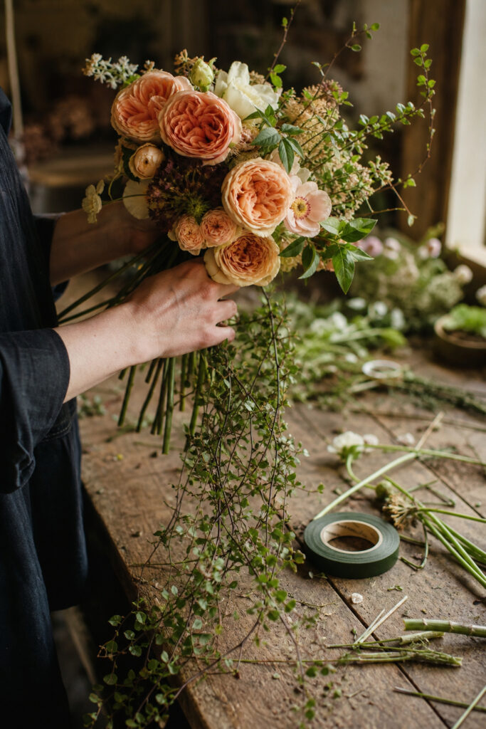 Hands assembling a boho cascade bouquet on a florist worktable with flowers and trailing vine