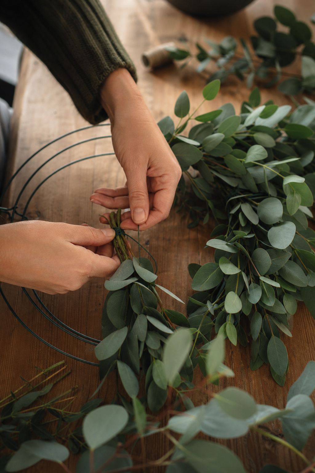 Hands wiring a small bunch of eucalyptus to a wire wreath frame with previous bunches overlapping