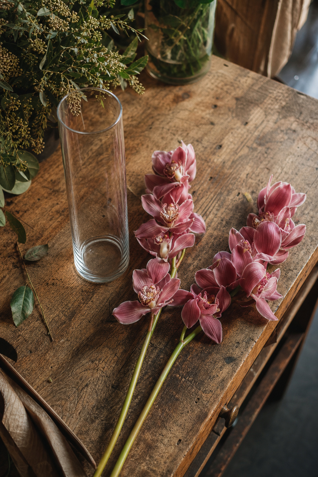 Empty tall narrow cylinder vase beside long cymbidium orchid sprays on a worktable