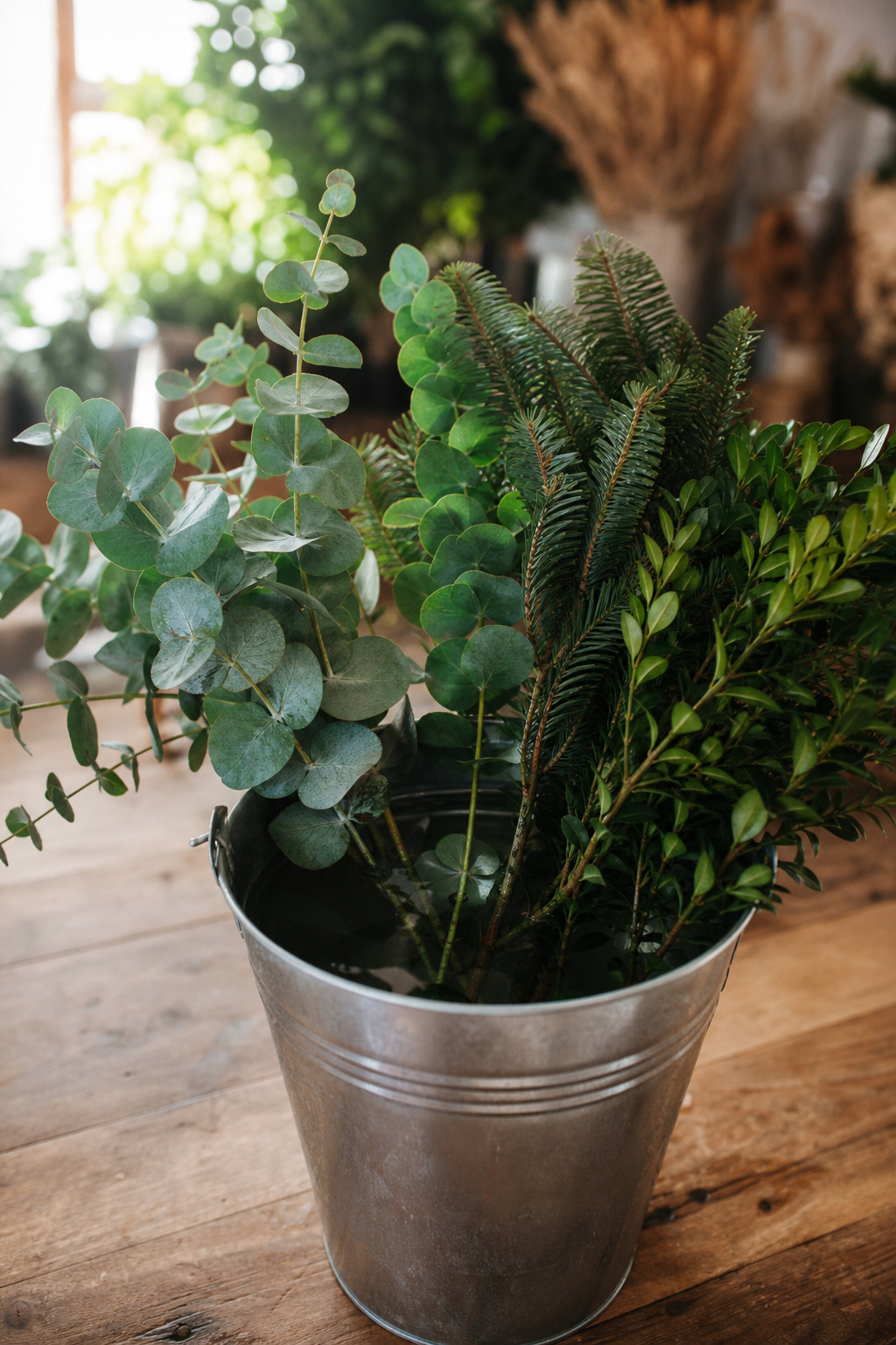 Eucalyptus, noble fir, and boxwood stems conditioning in a bucket of water on a florist's worktable