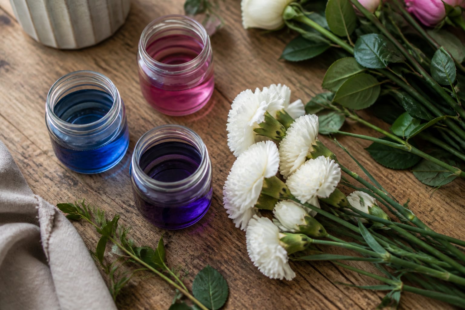 Three jars of floral dye solution beside white flowers on a florist's worktable