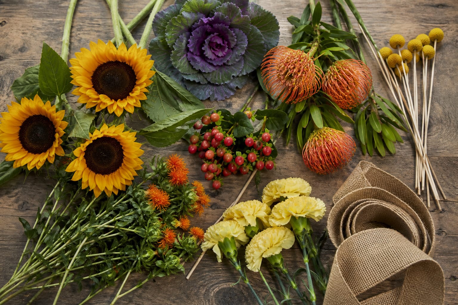 Fall flowers and floral supplies including sunflowers, protea, craspedia, and burlap ribbon laid out on a table
