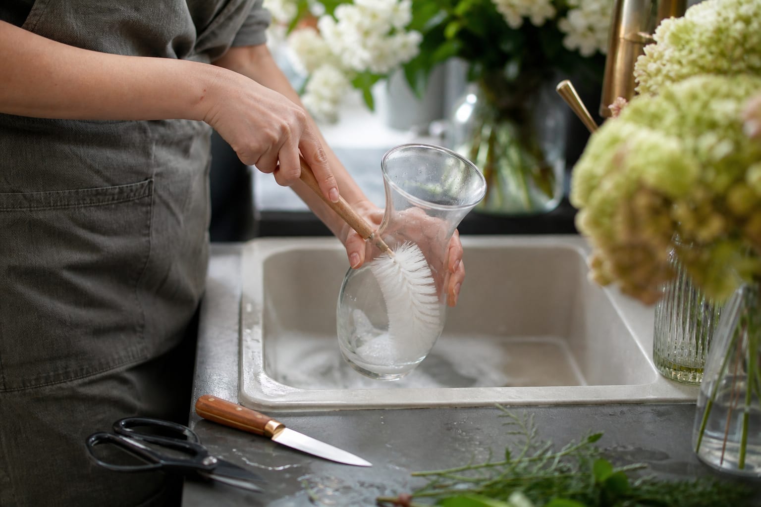 Florist washing a glass vase with soap and a bottle brush next to clean floral tools