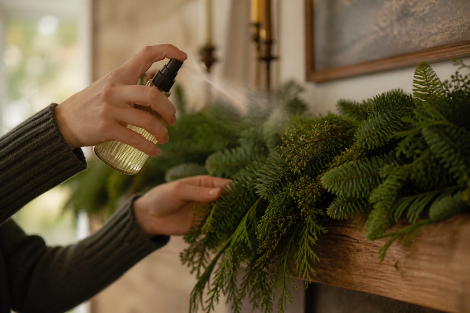 Hands misting a finished evergreen Christmas garland with a spray bottle on a mantel