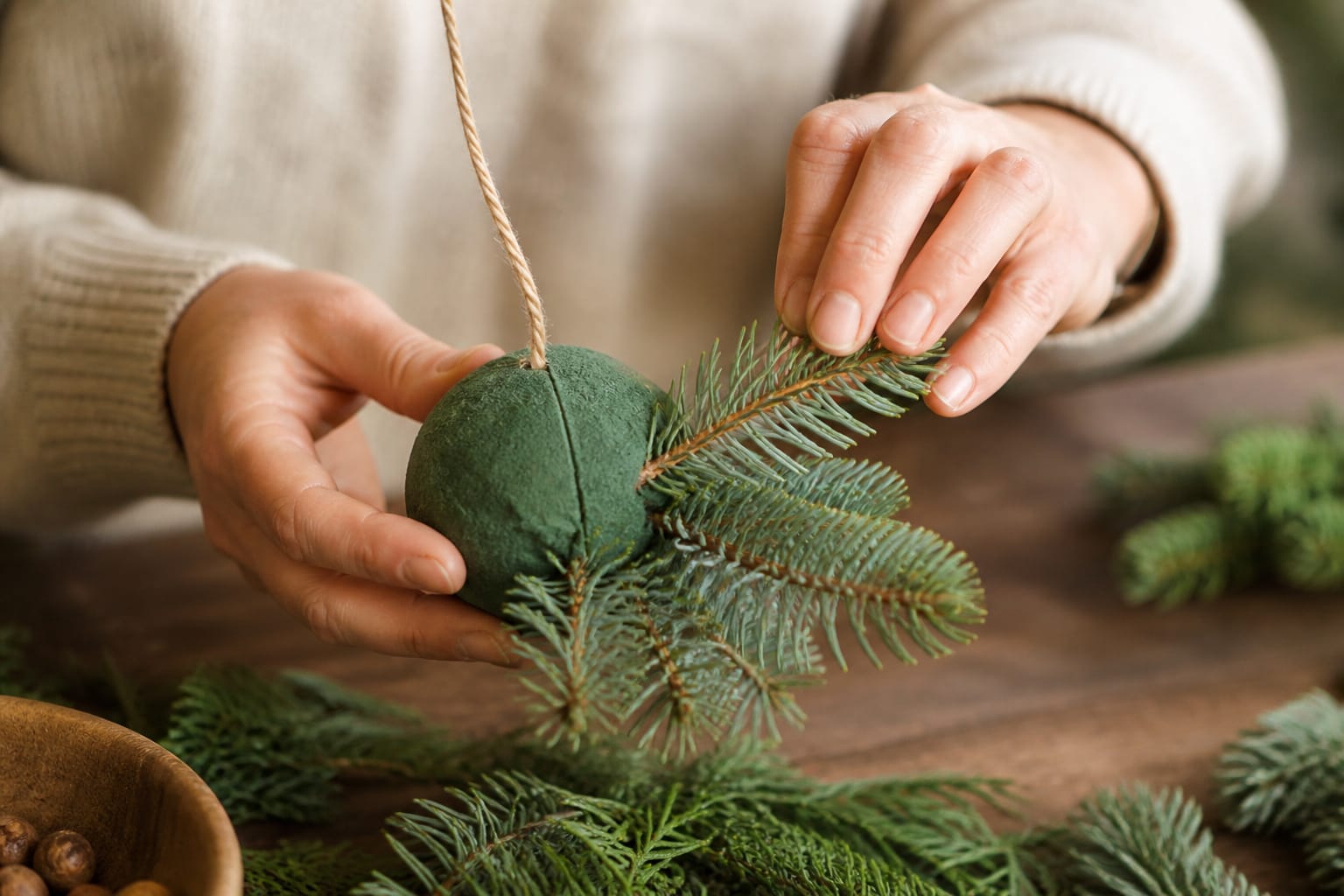 Hands assembling a Christmas kissing ball by inserting evergreen sprigs into a foam sphere