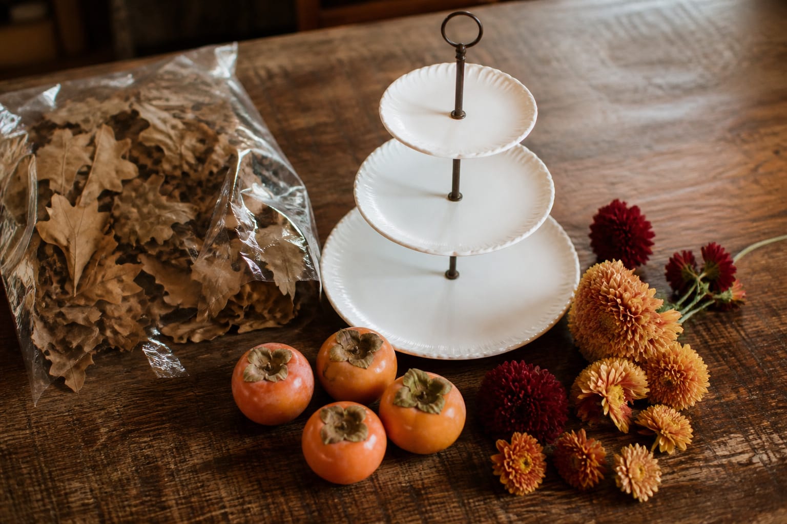 Supplies for a tiered floral centerpiece including a dessert stand, preserved foliage, persimmons, and mum heads