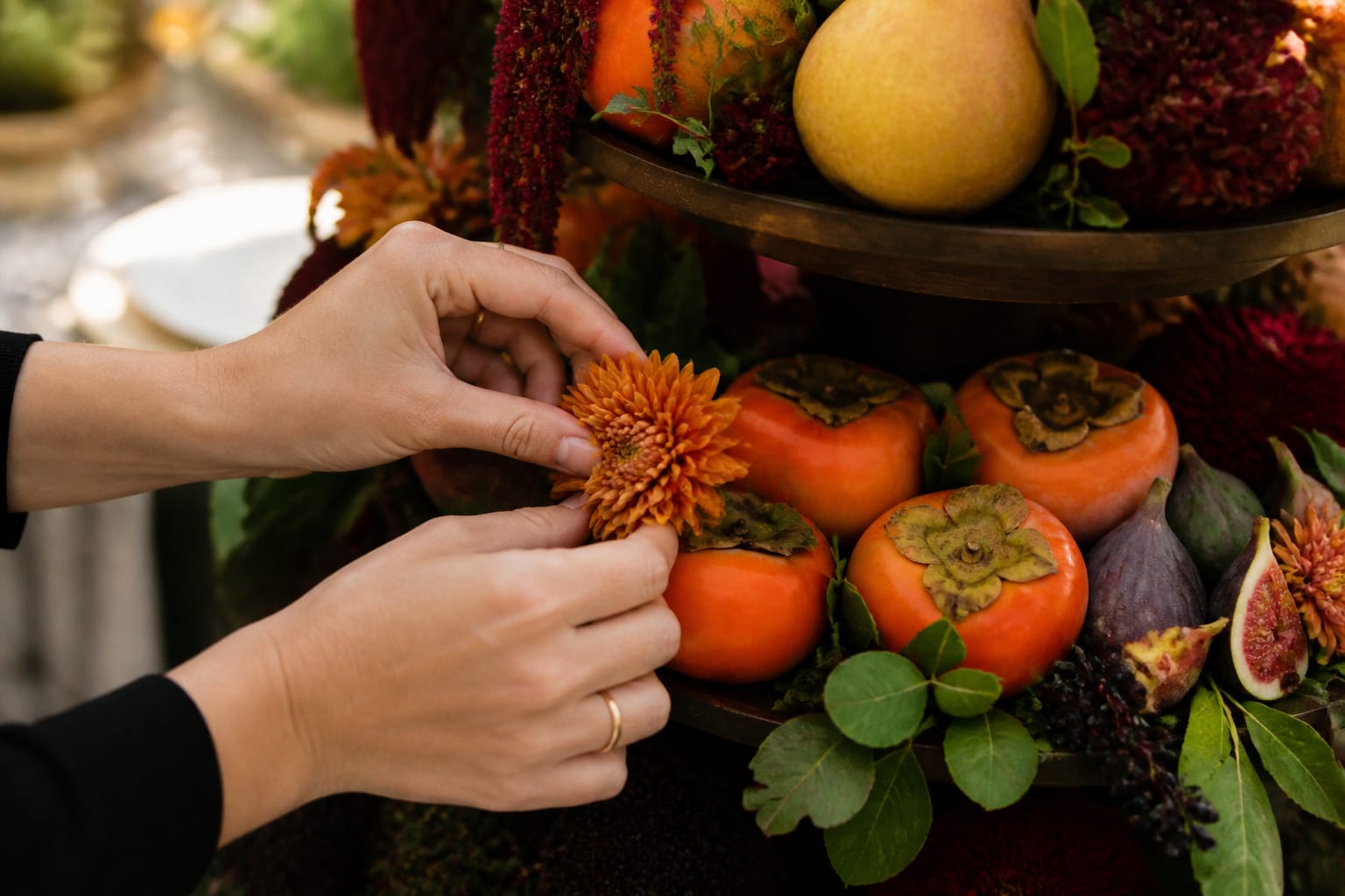Hands tucking a trimmed mum head between persimmons on a tiered centerpiece