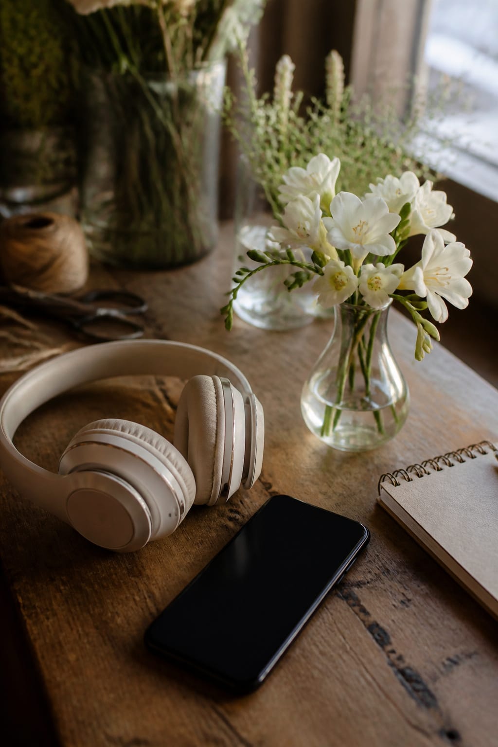 Over-ear headphones and a phone resting beside a small vase of flowers on a worktable